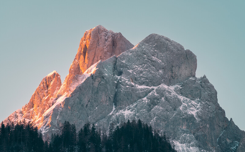 Snow-covered mountain peaks with golden alpenglow on the left face and dense evergreen forest at the base.