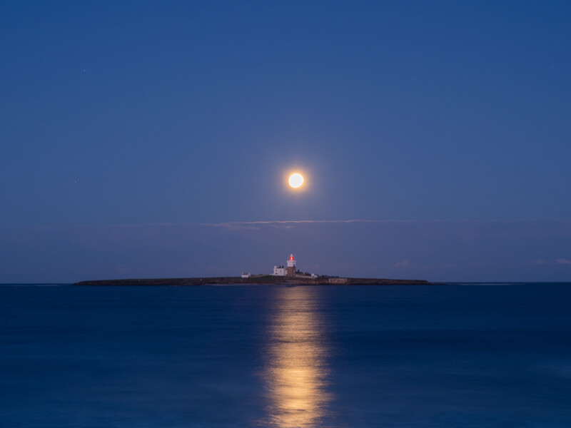 Full moon rising above a distant lighthouse on the horizon at twilight, with its reflection stretching across calm blue water.