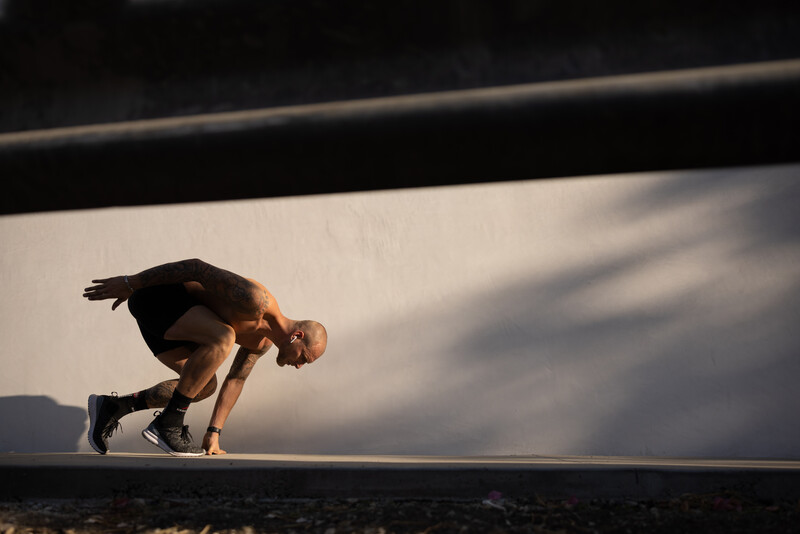 Shirtless athlete in a deep forward fold stretch position on a concrete platform, backlit by golden sunlight.