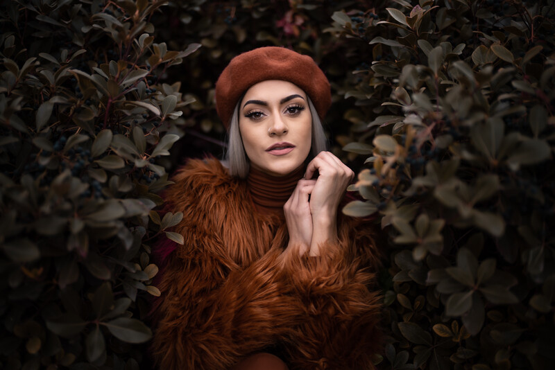 Woman with long reddish hair wearing a rust-colored beret and fuzzy jacket, posed among dark foliage.
