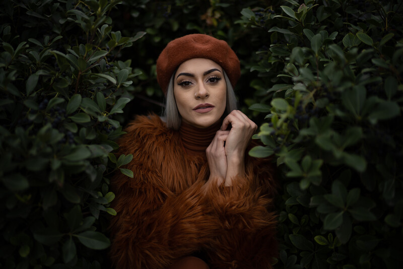Woman with pale complexion wearing a rust-colored beret and matching fuzzy jacket, surrounded by green foliage.