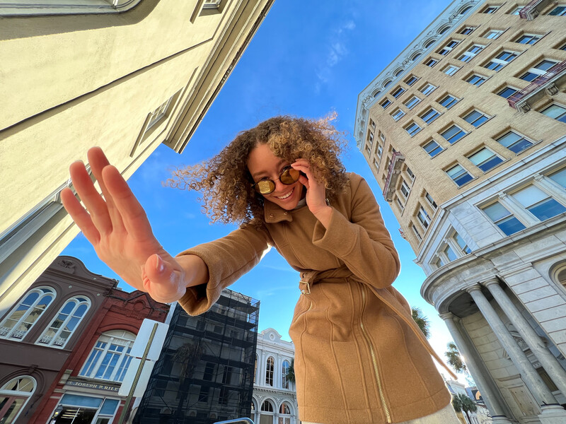 Person with curly hair and glasses in camel coat reaching toward camera with downtown buildings framing upward perspective.
