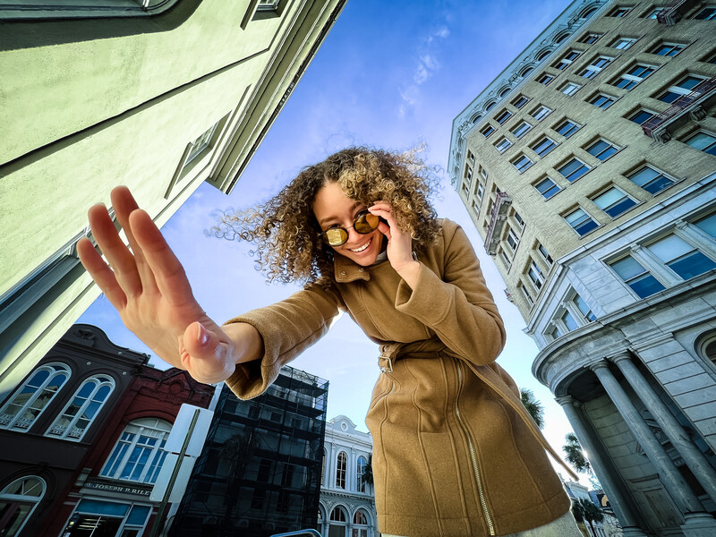 Woman in tan coat looking down at camera with hand raised, shot from below against urban high-rise buildings and blue sky.