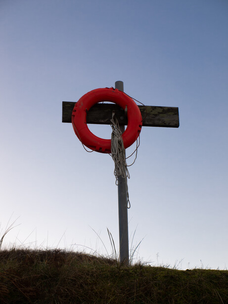 Wooden cross with bright red life ring mounted on grassy hillside against clear blue sky.