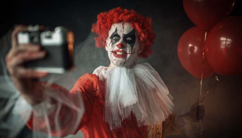 Clown in red and white costume holding red balloons while taking a selfie with a smartphone.
