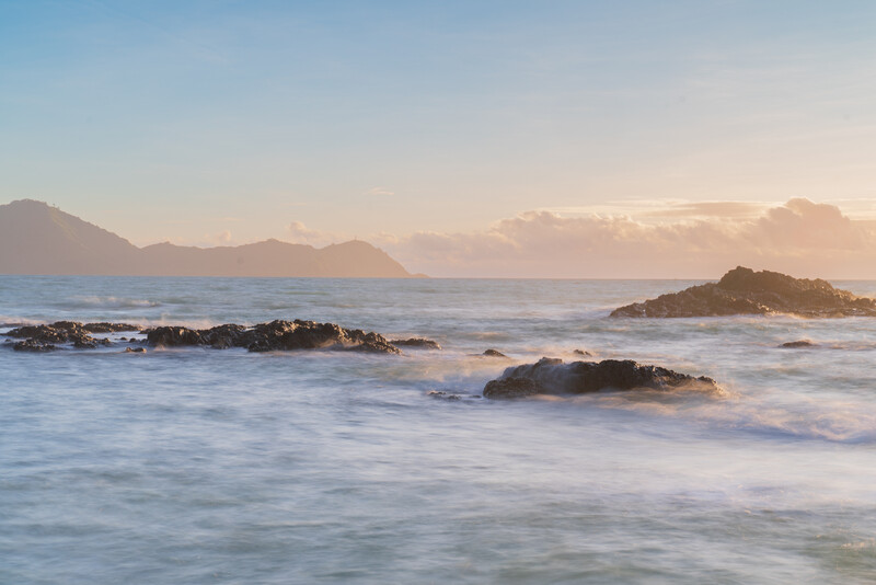 Rocky outcrops in shallow ocean waves at golden hour, with distant mountains under soft clouds.