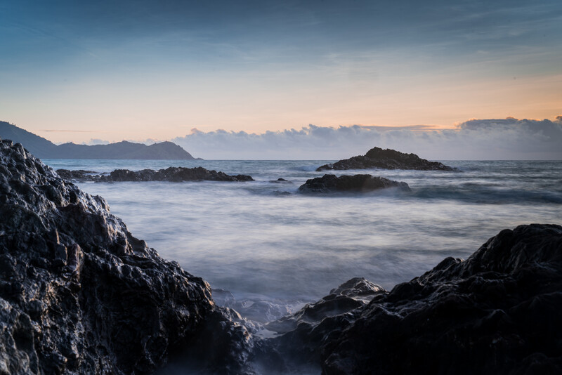 Rocky coastline at sunrise with silhouetted islands and long-exposure waves under cloudy sky.