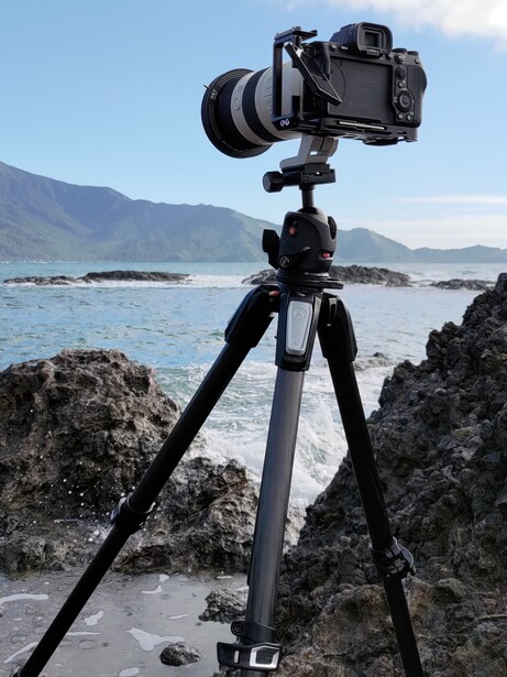 Professional camera on tripod mounted on rocky coastal shoreline with ocean and mountains in background.