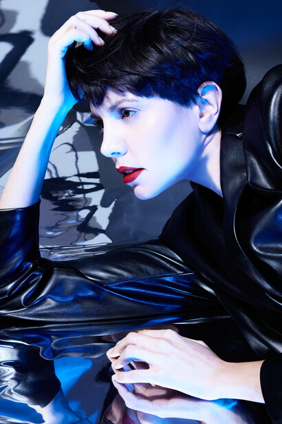 Person with short dark hair and red lipstick posed against reflective blue surfaces in studio lighting.