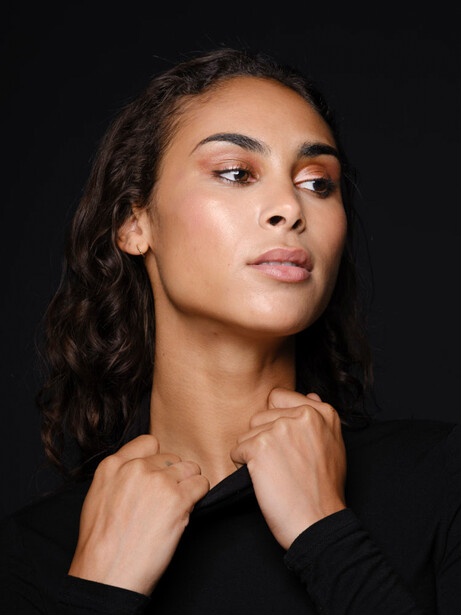 Woman with long dark hair gazing upward against a black background, hands near her neck.