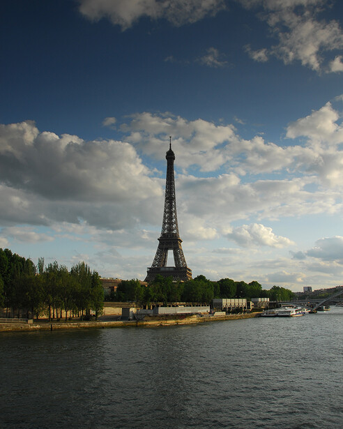 Eiffel Tower rising from the Seine riverbank under a partly cloudy sky with riverside buildings and boats visible.