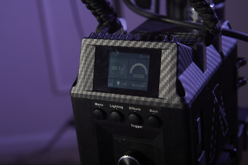 Professional lighting control monitor with carbon fiber housing, displaying timecode and settings against purple studio backdrop.