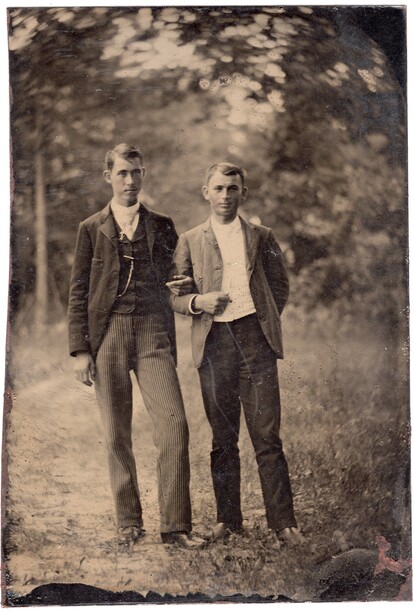Two men standing outdoors on a tree-lined path, wearing early 20th-century suits and hats.