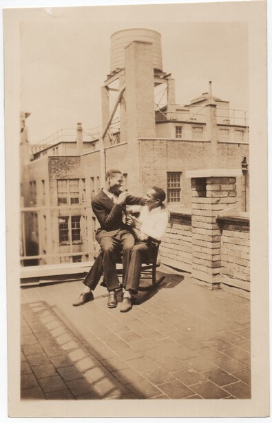 Man seated on rooftop holding camera, industrial brick buildings with water tower visible behind.