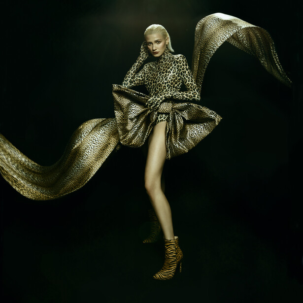 Model suspended against dark background wearing gold patterned garment with dramatic flowing fabric.
