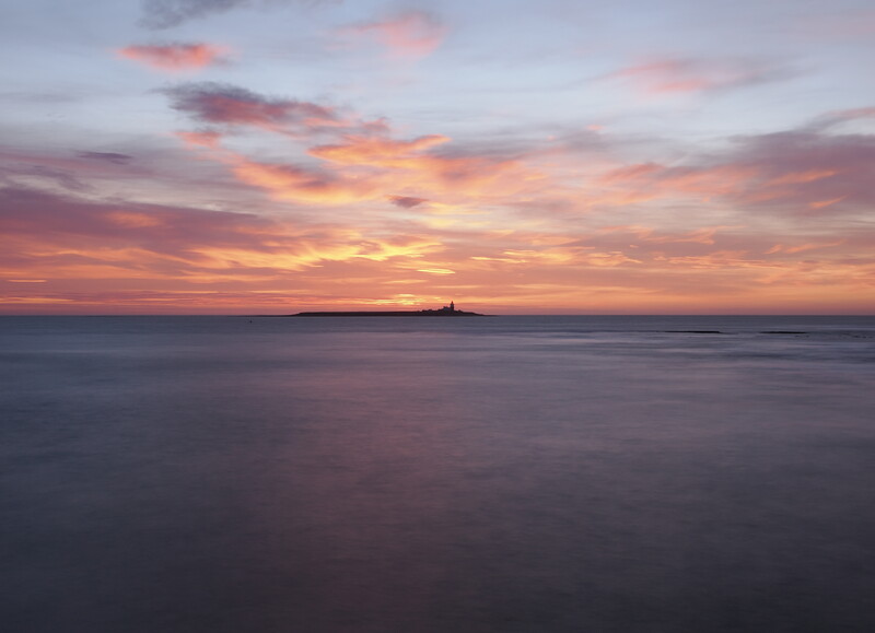 Sunset over calm water with a distant silhouetted island on the horizon.