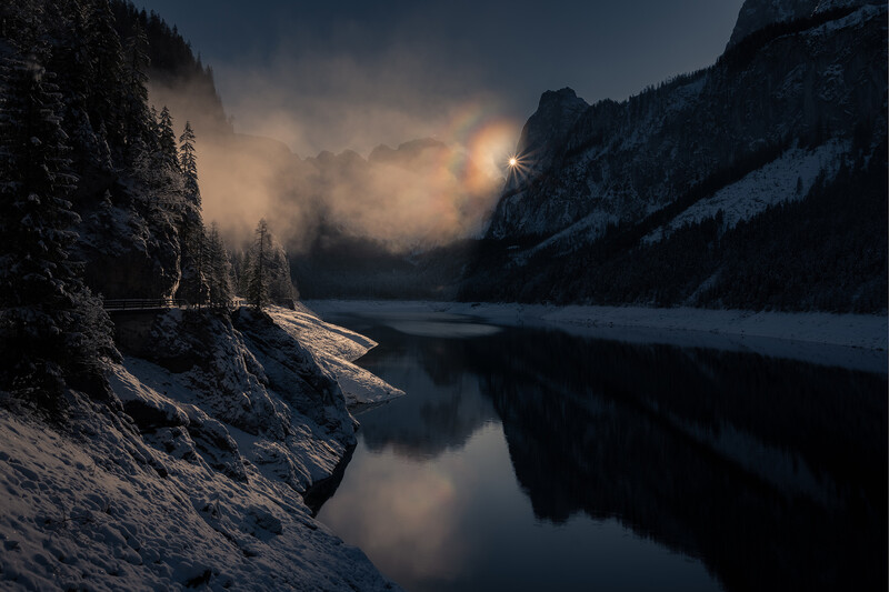 Snow-covered mountain valley with frozen lake reflecting dramatic clouded sky and distant sun.