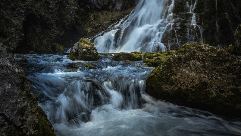 Cascading waterfall flowing over moss-covered rocks in a narrow canyon.