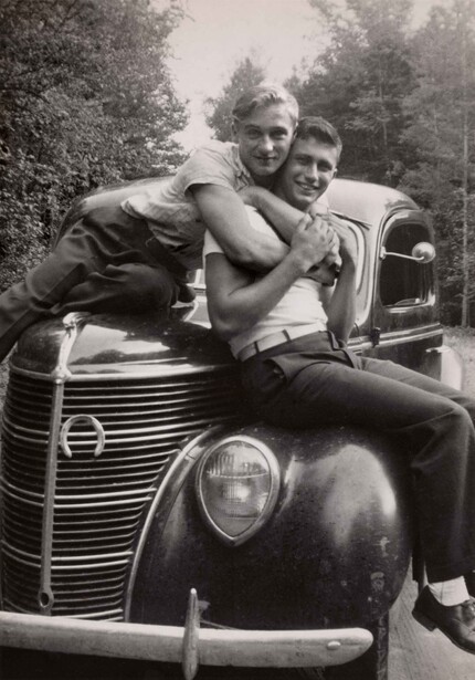 Two men posing together on the hood of a vintage 1930s automobile in black and white.