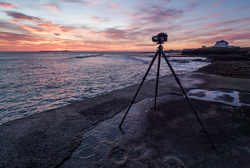 Camera on tripod positioned on rocky beach during colorful sunset with pink and orange sky.