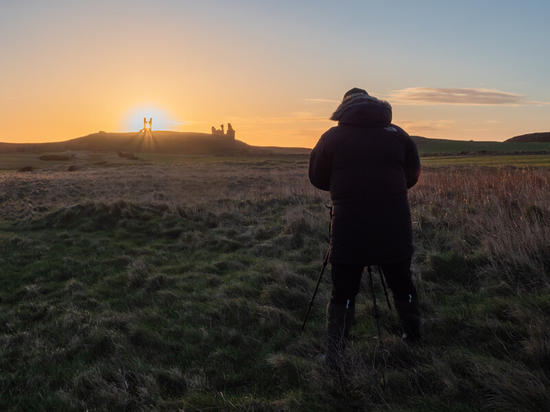 Photographer with tripod silhouetted against golden sunset over distant ruins on horizon.