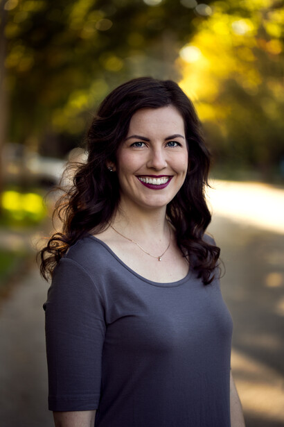Woman with long dark wavy hair and red lipstick smiling outdoors in natural light.