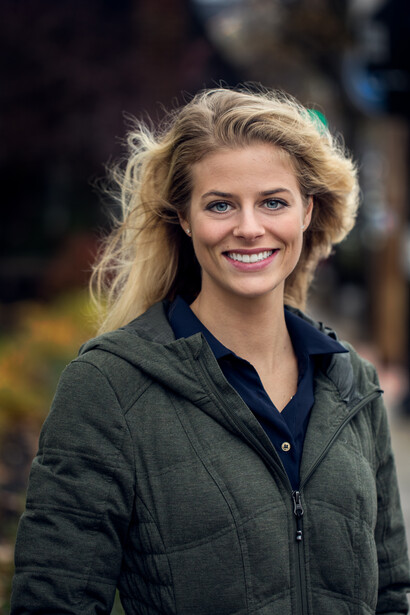 Woman with blonde hair smiling at camera wearing a dark quilted jacket outdoors.