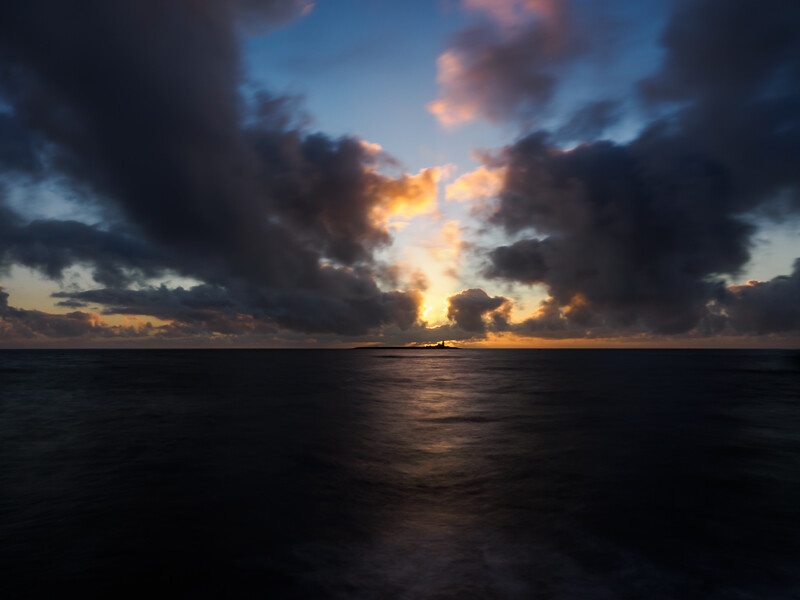 Dramatic sunset over calm ocean with golden light breaking through dark storm clouds.