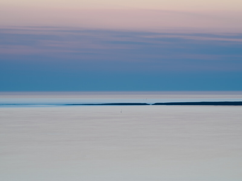 Minimalist seascape with soft pink sky meeting calm blue water and pale foreground under twilight conditions.