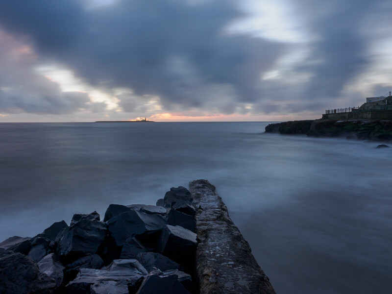 Long exposure seascape with rocky breakwater extending into calm water at dusk.