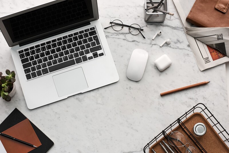 Flat lay of a white laptop, wireless mouse, earbuds, eyeglasses, brown leather wallet, notebook, pen, and small potted plant on a marble desktop.