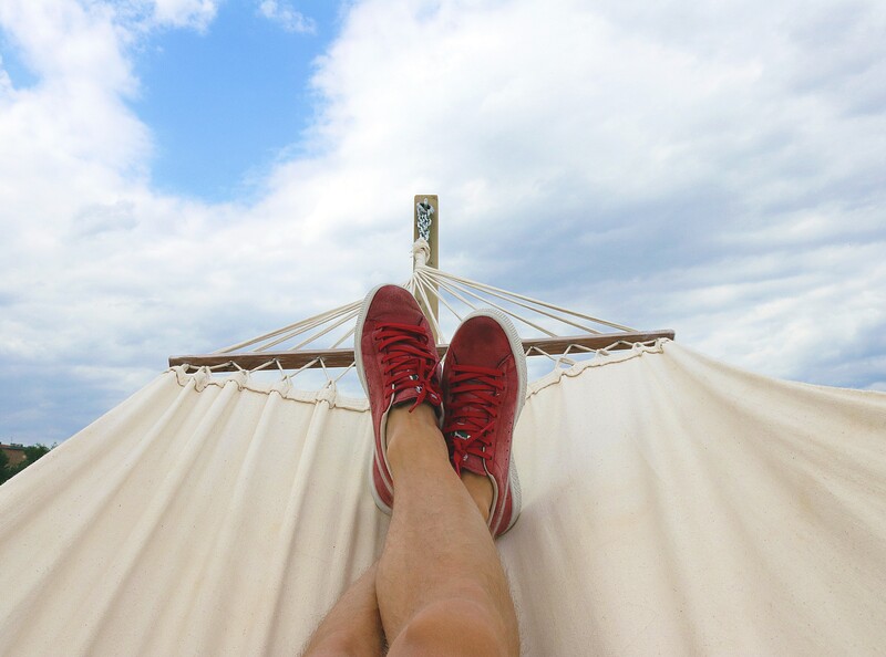Person's legs in red shoes resting on canvas hammock with bridge structure visible above against cloudy sky.