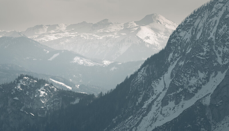 Snow-covered mountain peaks rise above a forested valley shrouded in mist and atmospheric haze.