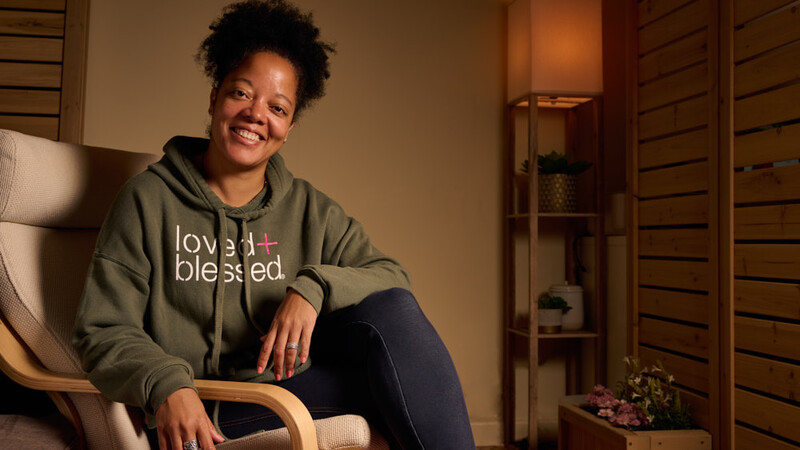 Woman in olive hoodie sitting in wooden chair, smiling at camera in warm studio setting.