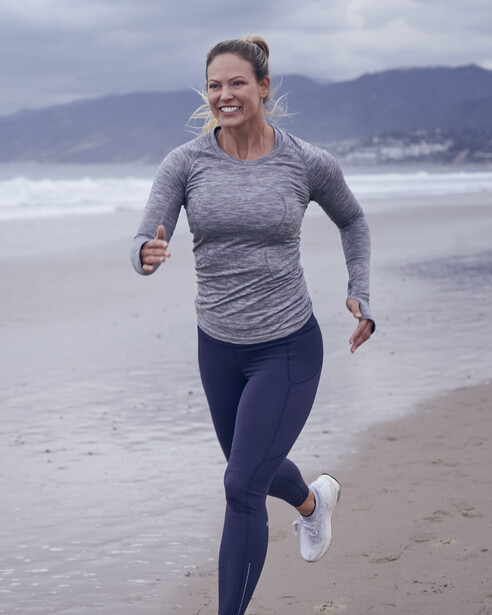 Woman jogging on a beach with mountains visible in the distance under overcast skies.