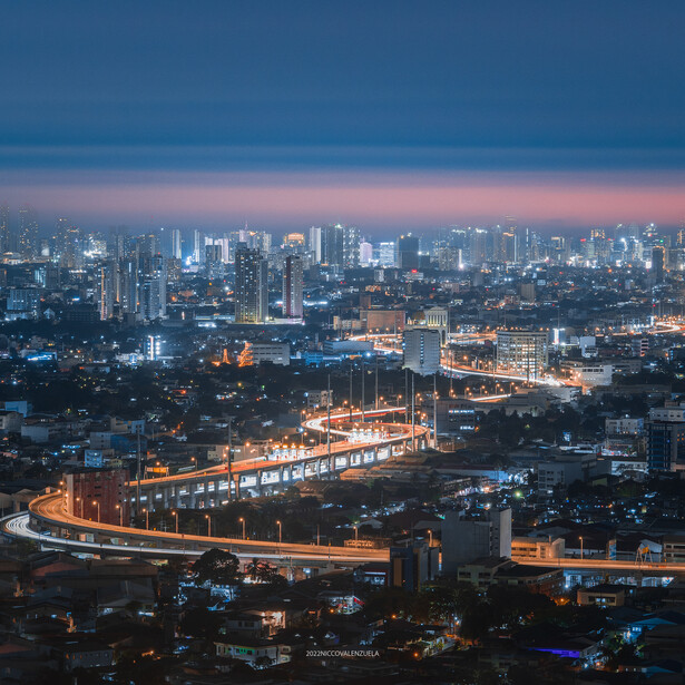 Illuminated elevated highway curving through a sprawling city at dusk, with downtown skyline glowing against twilight sky.
