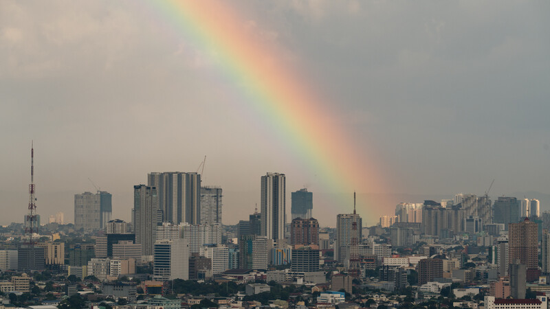 Vivid rainbow arcing over a dense urban skyline with high-rise buildings and communication towers.