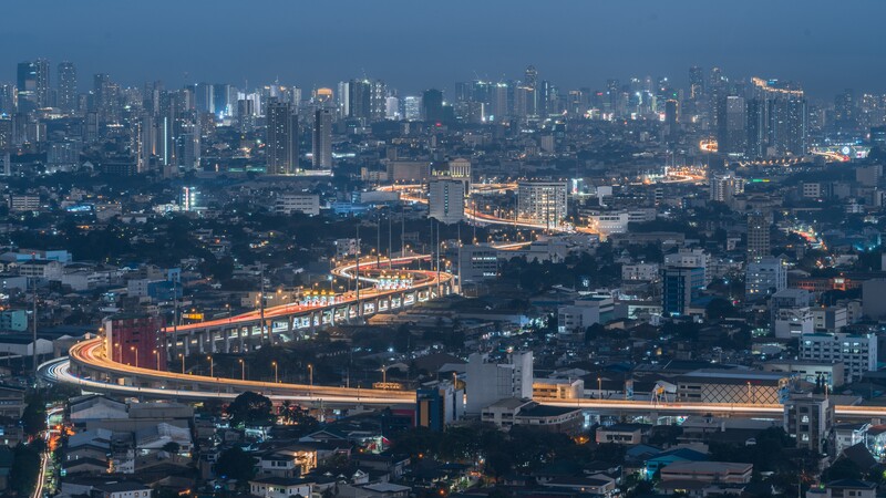 Illuminated elevated highway curving through a dense urban cityscape at dusk with skyscrapers in the background.