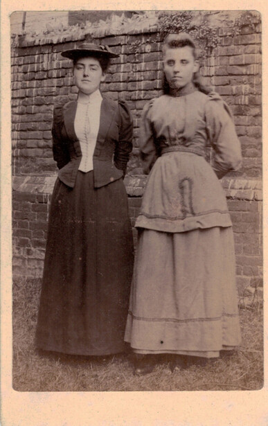 Two women in long dresses standing against a brick wall, circa early 1900s.