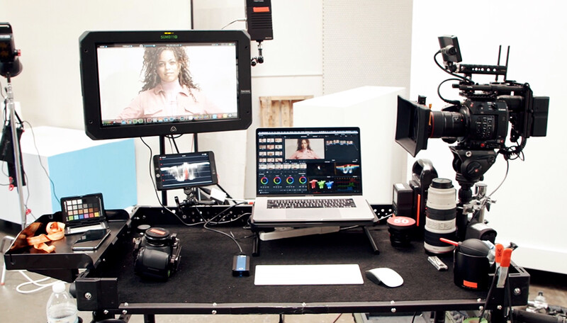 Professional video production workspace with monitor displaying talent, editing laptop, camera rig, and lighting equipment.