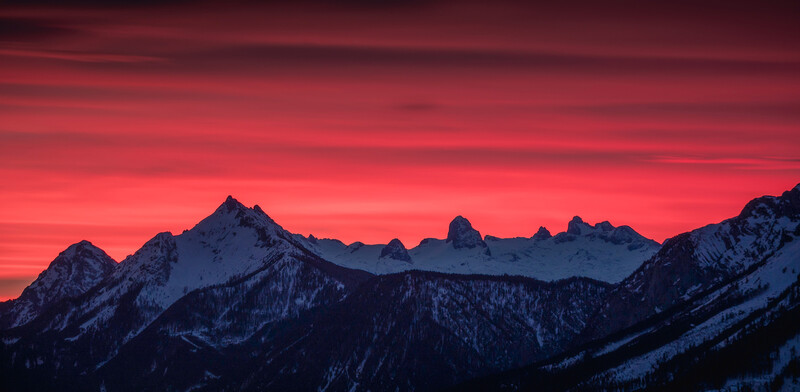 Snow-covered mountain range silhouetted against a vivid red and pink sunset sky.