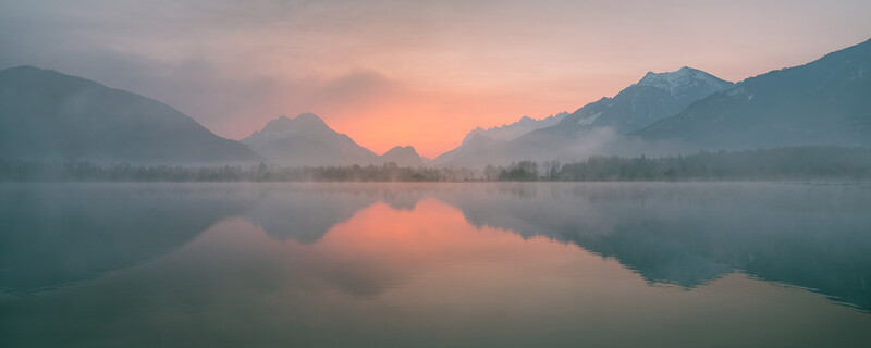 Mountain lake at sunrise with mirror-like reflections and mist rising from the water surface.