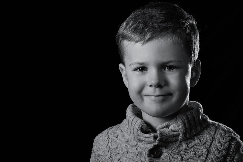 Young boy with short blonde hair wearing a cable-knit sweater, smiling at camera against black background.