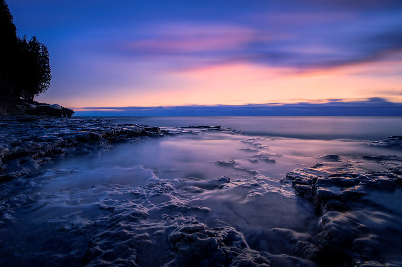 Rocky shoreline at twilight with calm water and purple-pink sky at dusk.