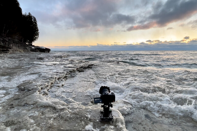Camera mounted on tripod in shallow ocean waves at sunset with rocky coastline and cloudy sky.