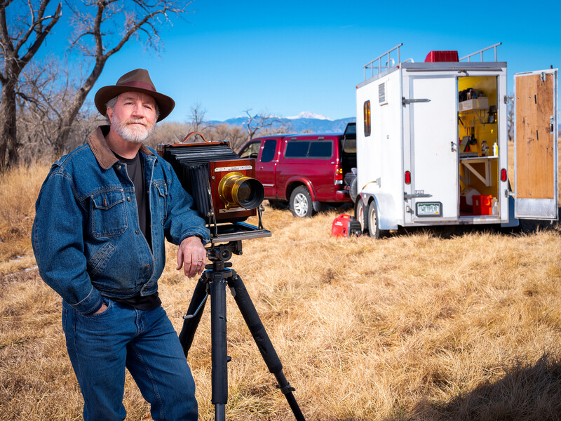Photographer operating a camera on tripod in desert landscape with vehicles and mountains.