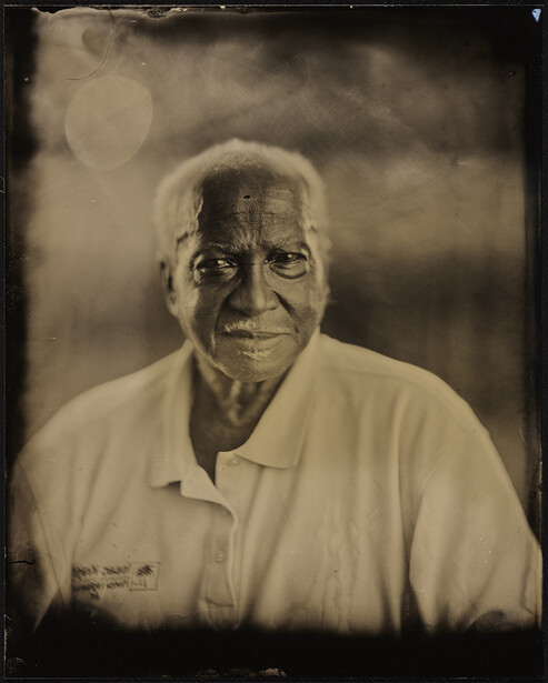 Portrait of an elderly man with white hair wearing a light-colored shirt, photographed against a soft, atmospheric background.