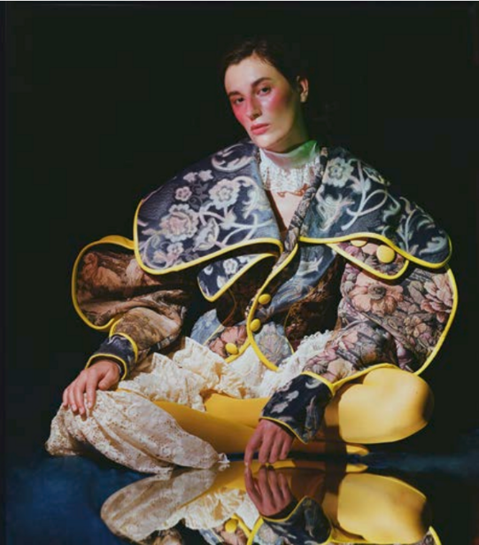 Person wearing patterned jacket surrounded by yellow lemons and white flowers against dark background.