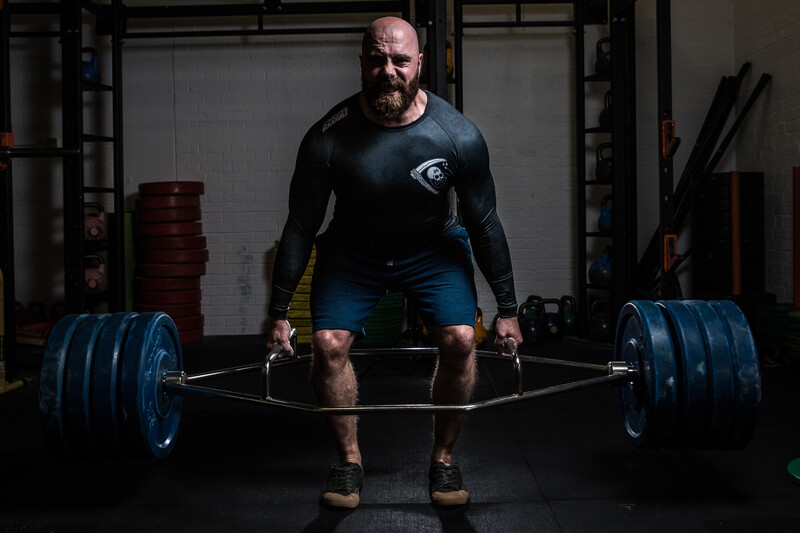 Bearded man in blue compression shirt performing a barbell deadlift with blue-plated weights in a gym.