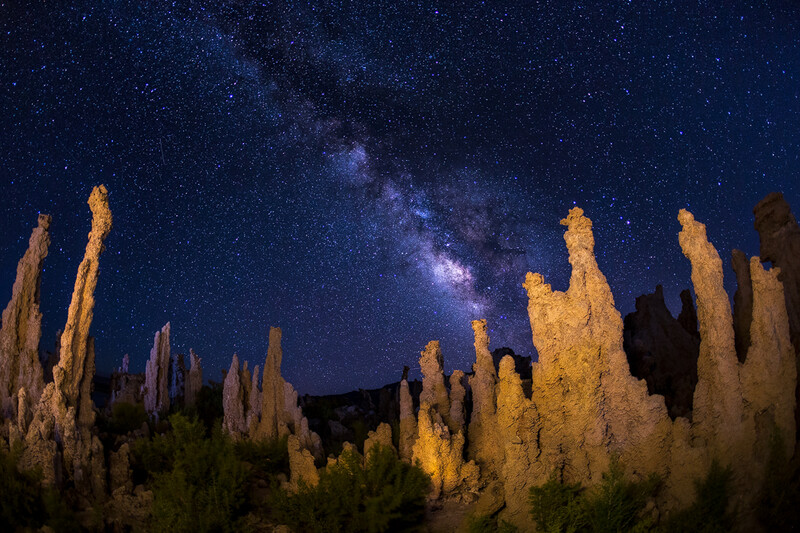 Milky Way galaxy stretches across night sky above illuminated desert rock formations.